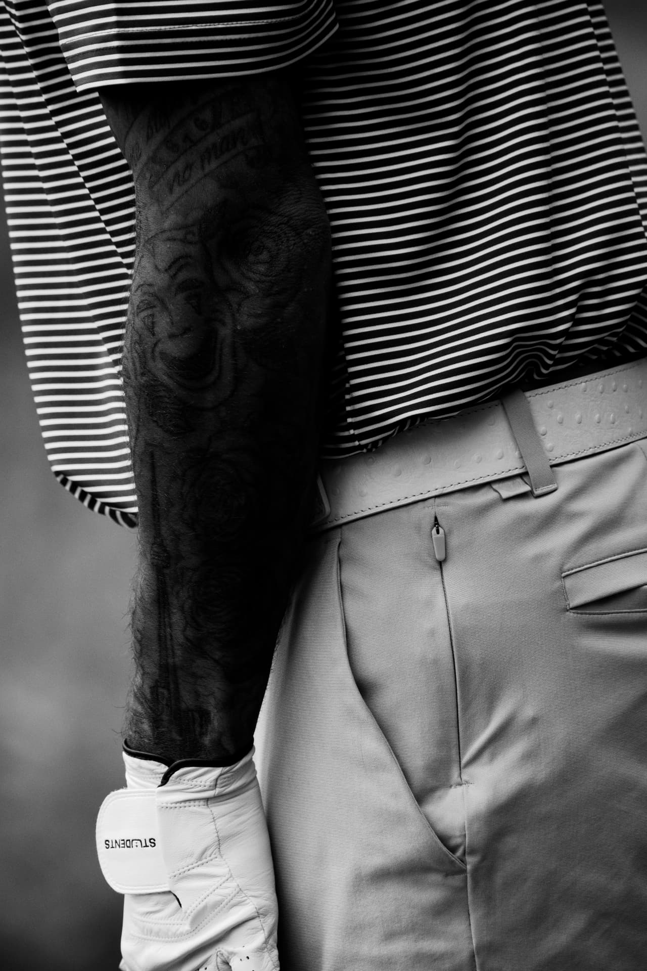 Black-and-white detail of a golfer in a striped polo and tailored trousers wearing a white Dartee ostrich belt